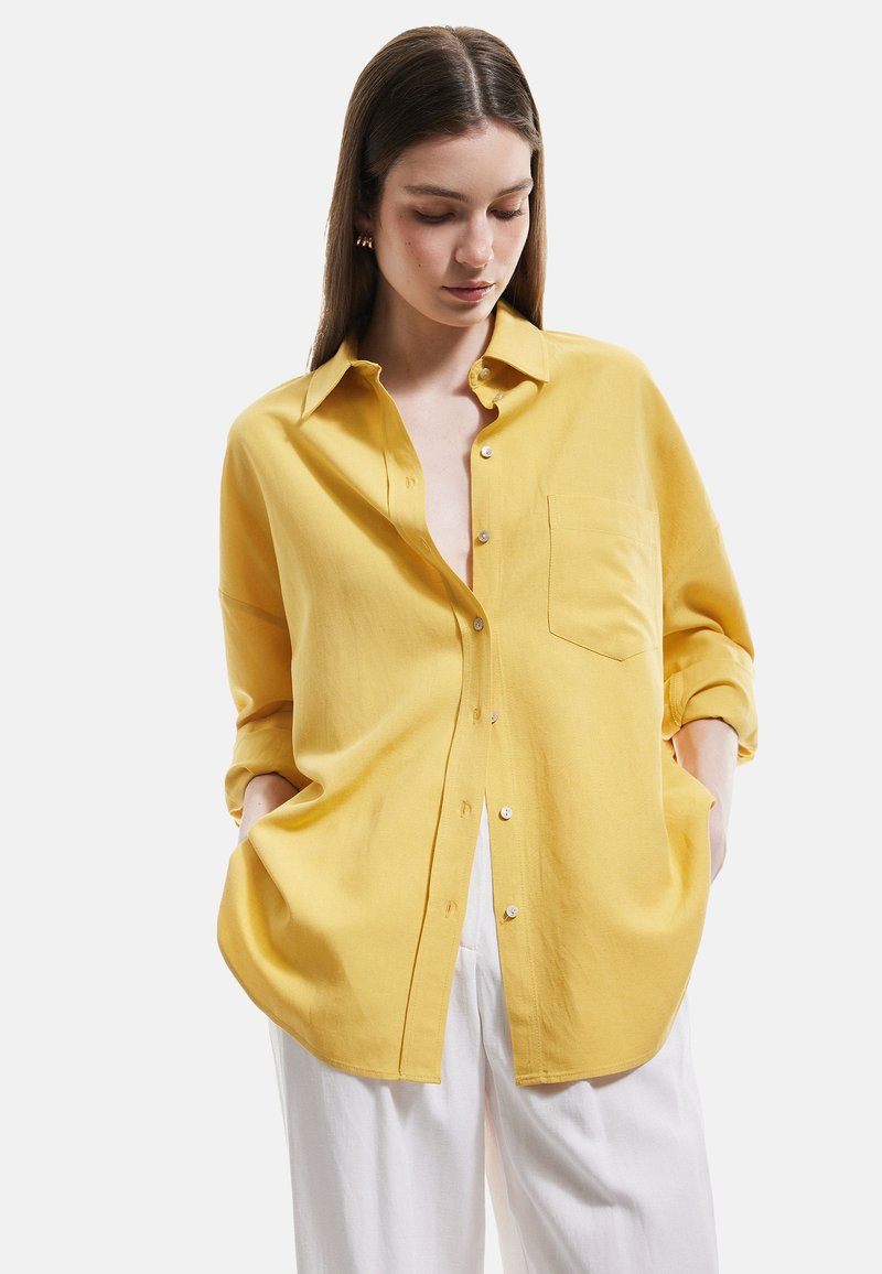 Gul button-up skjorte lavet af blødt stof, med krave, en brystlomme og opstegne ærmer. Sammen med hvide bukser.