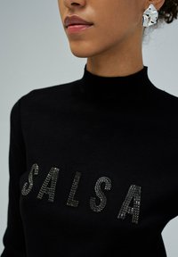 Kobieta ubrana w czarny sweter typu golf z napisem "SALSA" wyszytym czarnymi cekinami oraz srebrnymi kolczykami w kształcie abstrakcyjnym.
