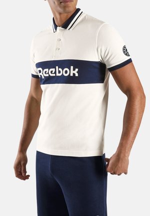 Hombre vistiendo una camiseta polo blanca con una franja horizontal azul marino y el logo de Reebok, combinada con pantalones azul marino.