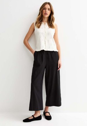 Jonge vrouw met los golvend haar, een mouwloos wit ooglet-topje met knopen, zwarte wijd uitlopende broek en zwarte Mary Jane schoenen, staand.