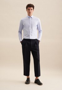 Lichtblauw overhemd met kraag, gecombineerd met donker, cropped op maat gemaakte broeken en zwarte veterschoenen. Effen achtergrond.