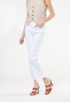 Beige mouwloos vest met knopen, witte cropped jeans met franjes aan de onderkant en bruine sandaaltjes met hak, enkelbandje en gespsluiting.