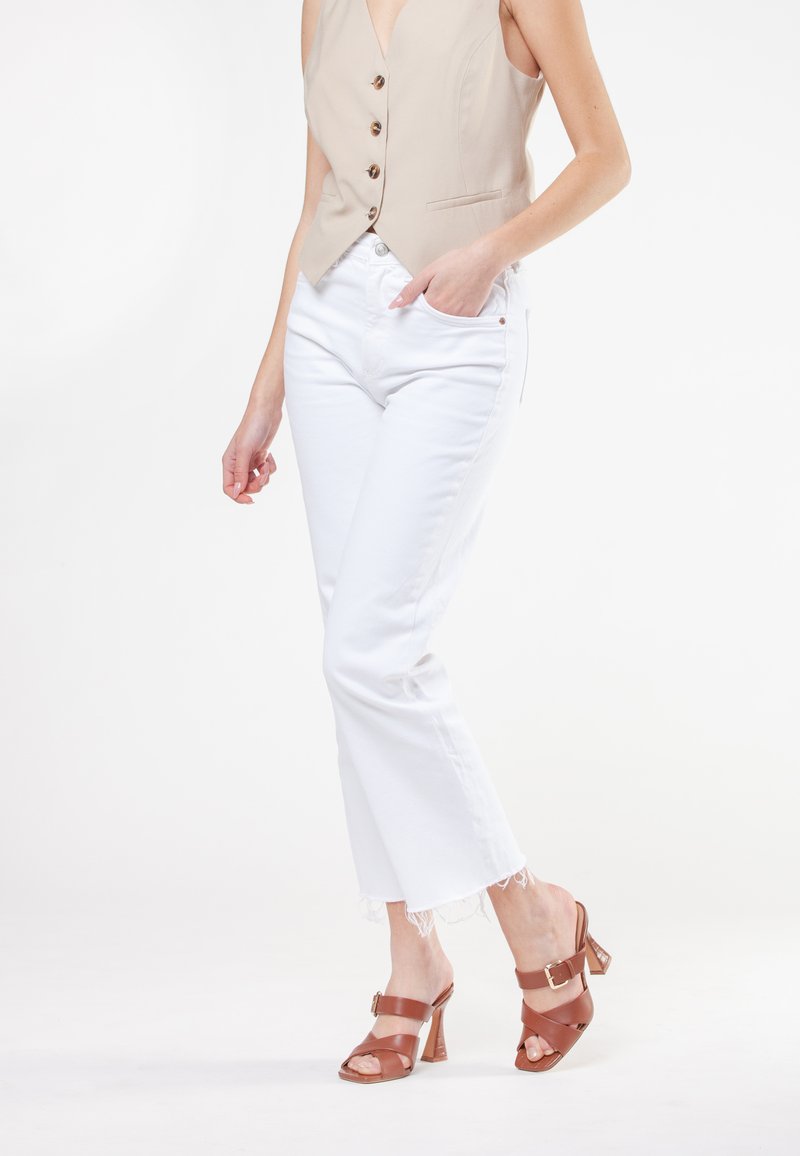 Beige ærmeløs vest med knapper, hvide cropped jeans med frynset kant og brune hælede sandaler med ankelrem og spænde detaljer.
