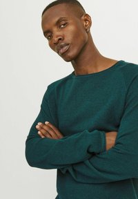 Mörk teal stickad tröja med rund halsringning, texturerat tyg och ribbade ärmslut. Modellen står med armarna korsade och visar upp passformen och designen.