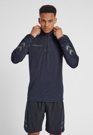 Maglietta sportiva a maniche lunghe navy con zip a un quarto e logo grigio sul petto. Presenta motivi a chevron grigi sulle maniche, abbinata a pantaloni corti neri.