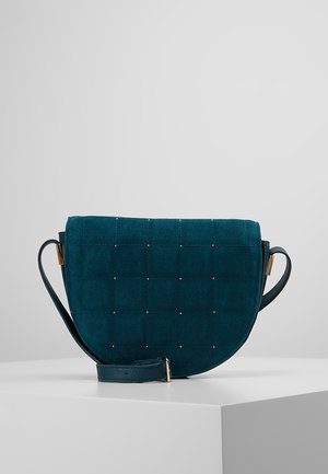 Tumma teal-värinen mokkanahkainen crossbody-laukku, jossa tikattu kuvio, pienet metallipidikkeet, läppäkiinnitys ja säädettävä nahkahihna valkoisella esittelytasolla.