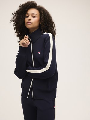 Mujer joven con cabello rizado que lleva una chaqueta y pantalones de chándal azul marino con franjas blancas y un pequeño logo en el pecho y el muslo.