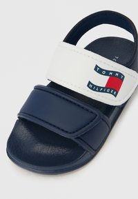 Sandalias azul marino con una amplia correa blanca que presenta el logo de "Tommy Hilfiger" y un diseño gráfico en rojo y azul. Suela texturizada.