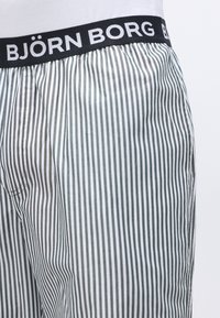 Melna josta ar baltu "BJÖRN BORG" tekstu uz baltām un pelēkām vertikālām svītrainām šortiem, ko valkā ar baltu kreklu.