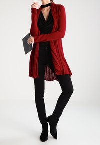Rote gestrickte Strickjacke mit langen Ärmeln und offener Vorderseite, kombiniert mit einem schwarzen Rollkragenoberteil und schwarzen schmal geschnittenen Hosen. Schwarze Knöchelstiefel.