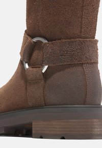 Hnědý semišový kotníkový bot s kulatým stříbrným kovovým detailem na texturovaném pásku a kontrastní tmavě hnědou gumovou podrážkou.