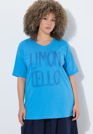 Mujer con cabello rizado que lleva una camiseta azul brillante con el texto "LIMONCELLO" en letras estilizadas, posando frente a un fondo blanco.