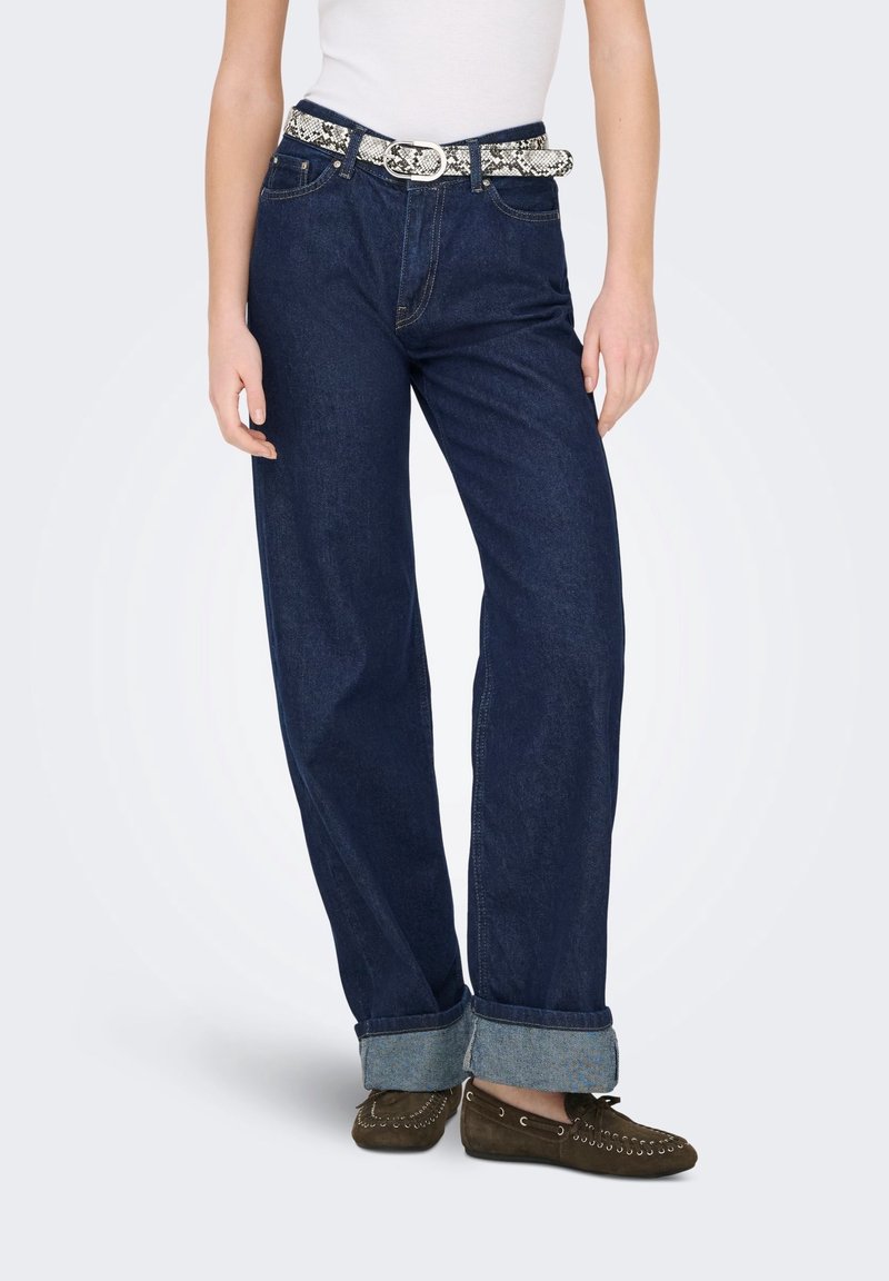 Femme portant un jean large bleu foncé avec des revers roulés, un top blanc, une ceinture imprimé serpent et des chaussures marron style mocassins.