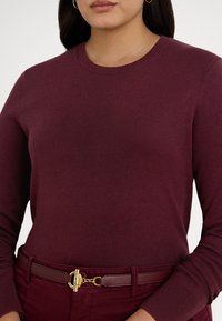 Γυναίκα που φοράει εφαρμοστό μπορντό πουλόβερ μακρυμάνικο και παντελόνι που ταιριάζει, με ζώνη με χρυσή αγκράφα, ορατή από το στήθος μέχρι τους γοφούς.