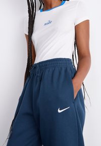 Bílé přiléhavé tričko s modrými detaily a nápisem „nike“ v modrém stehu, doplněné tmavě modrými tepláky se šňůrkou a bílým logem.