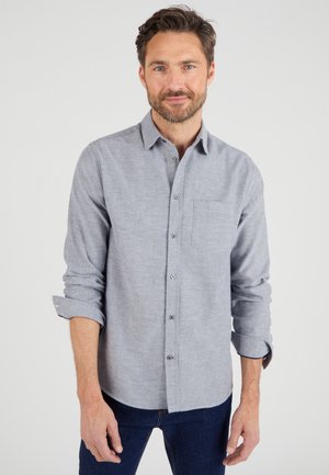 Camisa de botones gris con un bolsillo, hecha de una tela texturizada. Presenta mangas enrolladas y botones plateados, combinada con unos jeans azul oscuro.