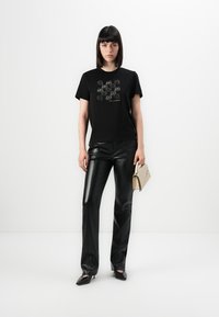 Schwarzes Baumwoll-T-Shirt mit Graf print, kombiniert mit glänzenden schwarzen Kunstlederhosen. Das Modell hält eine beigefarbene Handtasche.