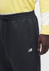 Detailný záber čiernych teplákov s elastickým pásom, šnúrkou, bočným vreckom a bielym logom New Balance na osobe v žlto-bielom tričku s dlhým rukávom.