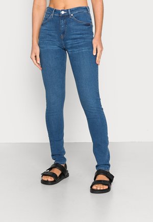 Kvinde iført tætsiddende blå jeans med høj talje og sorte åbne sandaletter, stående mod en ensartet lys baggrund.