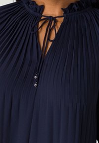 Haut bleu marine plissé avec un col à volants, noué avec un cordon en tissu et des accents de perles argentées. Tissu doux et texturé avec des plis verticaux.