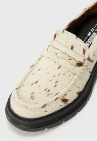 Kermavaaleat loaferit, jotka on valmistettu pehmeästä tekstuuri materiaalista, jossa on ruskeita laikkuja. Niissä on musta kumipohja ja virtaviivainen muotoilu.