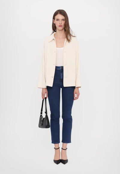 Vaalea beige takki valkoisen paidan päällä, yhdistettynä tummansinisiin slim fit -farkkuihin. Musta käsilaukku ja kärkitossut viimeistelevät asun.