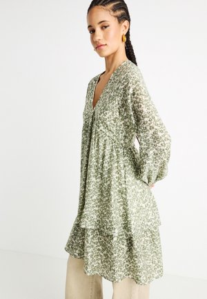 Νεαρή γυναίκα με πλεξούδες, φοράει ένα πράσινο και λευκό φόρεμα με λουλουδάτο σχέδιο και μακριά μανίκια, στέκεται μπροστά σε απλό λευκό φόντο.