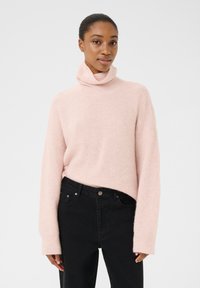 Svetro s turtleneck iz light roza barve, dolgimi rokavi, rebrasto teksturo in sproščenim krojem, v kombinaciji z črnimi kavbojkami. Brez vidnih vzorcev ali kovinskih delov.