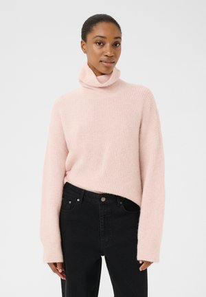Lyserød turtleneck sweater med lange ærmer, ribbet tekstur, afslappet pasform, parret med sorte jeans. Ingen synlige mønstre eller hardware.