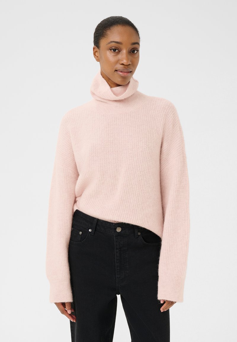 Svetro s turtleneck iz light roza barve, dolgimi rokavi, rebrasto teksturo in sproščenim krojem, v kombinaciji z črnimi kavbojkami. Brez vidnih vzorcev ali kovinskih delov.