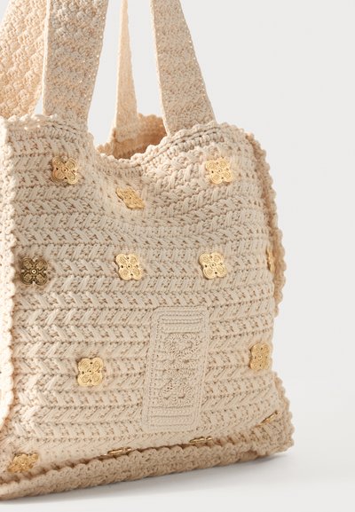 Τσάντα χειροποίητη με βελονάκι σε κρεμ χρώμα με χοντρούς ιμάντες και χρυσές διακοσμήσεις σε υφασμάτινη, υφή επιφάνεια.