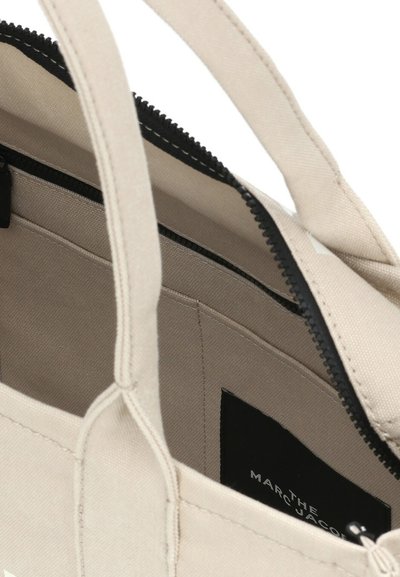 Bolsa tote beige com zíper e alças pretas. O interior apresenta um pocket e um tecido texturizado. Etiqueta com "THE MARC JACOBS" visível.