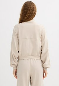 Sudadera beige de manga larga y corte corto con un ajuste relajado, que presenta una costura horizontal en la parte trasera y mangas fruncidas. Textura de tela suave.