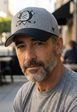 Middle-aged man with gray beard wearing a gray and black baseball cap with a hockey mask logo, dressed in a gray t-shirt, outdoors in daytime.
