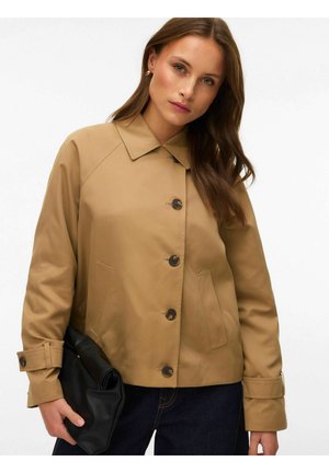 Femme aux longs cheveux bruns portant une veste beige boutonnée, tenant une pochette noire pliée, debout devant un fond clair uni.