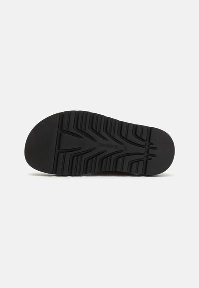 Semelle de chaussure en caoutchouc noir avec motif de bande de roulement texturé, comportant des rainures et des crêtes pour une meilleure adhérence. Détail de logo centré visible dans le design.