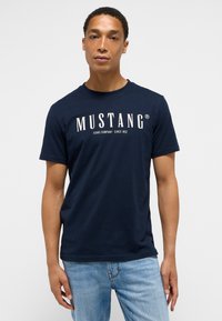 Tmavě modré tričko s krátkým rukávem vyrobené z bavlny, s bílým logem a textem "MUSTANG". Jednoduchý styl s kulatým výstřihem, hladká textura.