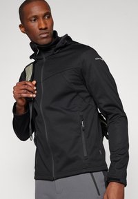 Giacca softshell nera con chiusura a zip, cappuccio regolabile, tasche laterali e una texture liscia. Presenta un branding discreto sulla spalla.
