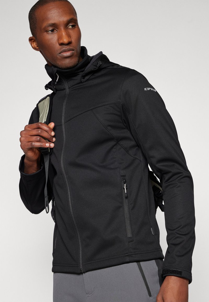 Schwarze Softshelljacke mit Reißverschluss, verstellbare Kapuze, Seitentaschen und glatter Oberfläche. Mit dezentem Branding auf der Schulter.