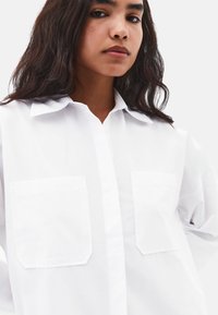 Weißes Langarmhemd mit Kragen. Verfügt über zwei Vorderseitentaschen und eine durchgehende Knopfleiste. Der Stoff wirkt glatt und leicht strukturiert.