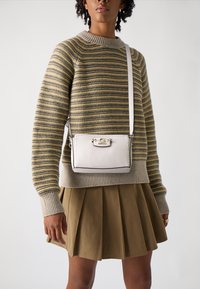 Borsa a tracolla in morbida pelle bianca con dettagli in argento, forma rettangolare, accentuata da cuciture, indossata con un maglione a righe e una gonna plissettata.