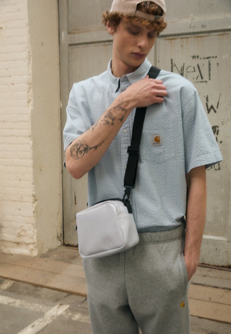 Sac en bandoulière blanc avec une texture lisse et une fermeture éclair, porté sur une chemise boutonnée texturée bleu clair et un pantalon de survêtement gris.