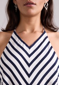 Navy ja valge triibuline ilma varrukateta tipp, millel on V-kaelus, chevron-muster ja sile kangatekstuur. Näha on hõbedased abstraktsed kõrvarõngad.