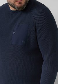 Navy gekleurde gebreide trui met een geribbeld ontwerp, ronde halslijn en een linkse borstzak met een drukknoopsluiting. Geaccentueerd met logo-detail.