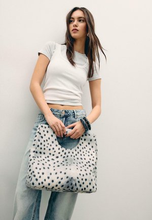 Young woman in light gray crop top and loose jeans holding large white handbag with black spots against plain background.