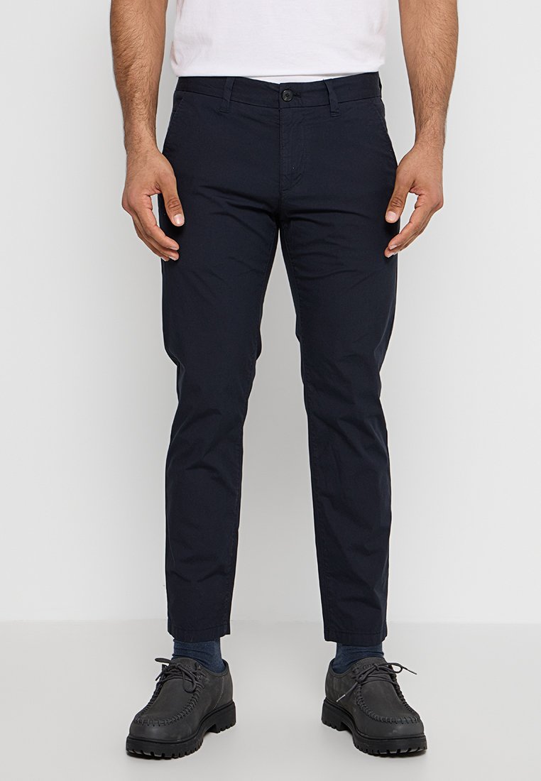 Mężczyzna w ciemnoniebieskich spodni slim fit, ciemnych skarpetach, czarnych butach na sznurówki i białym t-shircie, stojący na tle jednolitego jasnego tła.