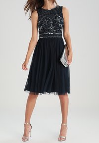 Cocktailklänning i mörkblått med en pärlbesatt, figursydd överdel och en flödande, knälång tylskjol. Inkluderar en silverclutch och klackskor med remmar.