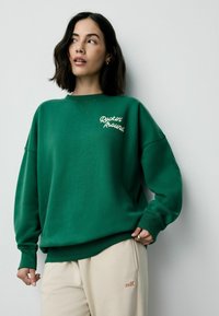 Grøn sweatshirt lavet af blødt stof, med "Rockin' Around" broderet i hvid, med en afslappet pasform og ribbede manchetter.