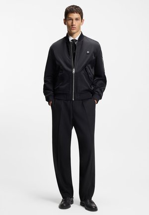 Mustasta, sileästä nylonkankaasta valmistettu bomber-takki, jossa on vetoketjusuljenta, resorit hihansuissa ja pieni logo. Yhdistetty räätälöityihin mustiin housuihin.