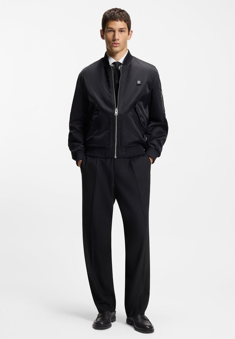 Zwart bomberjack gemaakt van glad nylon, met ritssluiting, geribde boorden en een klein logo. Gecombineerd met op maat gemaakte zwarte broek.